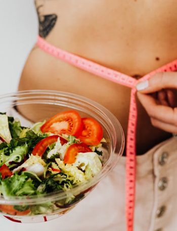 Woman holding vegetable salad and measuring waist with tape, symbolizing healthy lifestyle choices.