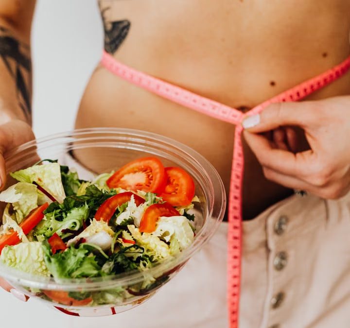 Woman holding vegetable salad and measuring waist with tape, symbolizing healthy lifestyle choices.